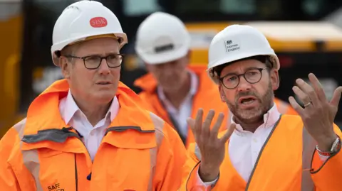 James Glossop/The Times/PA Wire Sir Kier Starmer facing another man, both are wearing orange high-vis jackets and white hard-hats. Starmer's hat has a red logo on the front that reads 'SISK'.