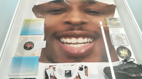 A photo of one of the exhibits with a large T-shirt with a man smiling on it, a vinyl, some CDs and some black trainers. 