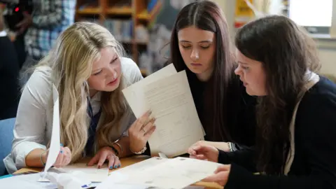 PA Media Three female school pupils receiving their exam results. They are sitting at a table looking at their certificates.