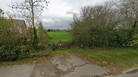 The metal gate at the entrance to fields which will make way for the new homes, with the outline of a house visible through the bushes to the side of the site