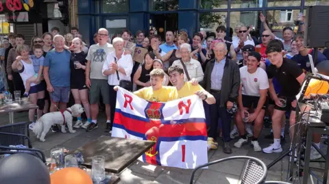 Helen Cowan The brothers crouch down holding an RNLI flag with a large crowd of people behind them cheering.