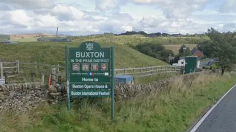 Green sign which reads "Buxton in the Peak District" in front of wooden fencing and green fields