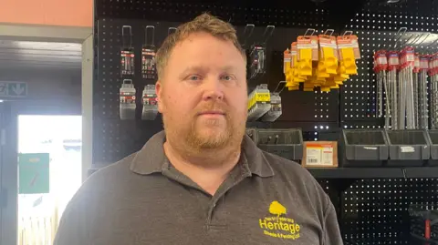 Shaun, who has ginger hair and a beard, is looking at the camera. He is wearing a grey polo with the yellow logo for his business on it, and there are tools visible behind him on racks. 