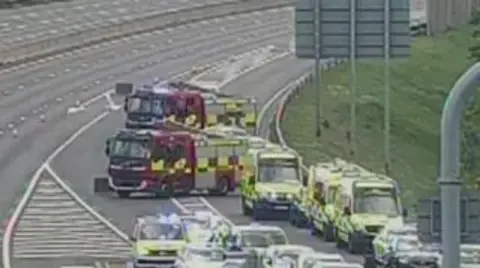 A grainy CC screenshot showing two fire engines and a several ambulances and a police car lined up on a motorway slip road.