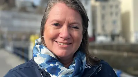 Christine Bangor-Jones is smiling at the camera. Her brown hair is swept back and she is wearing a light blue and white floral scarf with a dark blue coat. The buildings on the front of St Peter Port are visible in the background but blurred. 