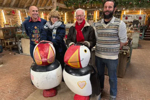 Inside the Repair Shop, standing behind the battered puffins before the makeover. Brendon wears a Christmas jumper, Dominic a patterned tank top and shirt, and Luise and Brian scarves and coats.