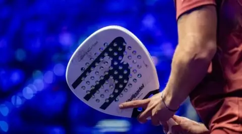 A man holding a white padel racquet with a blue background.