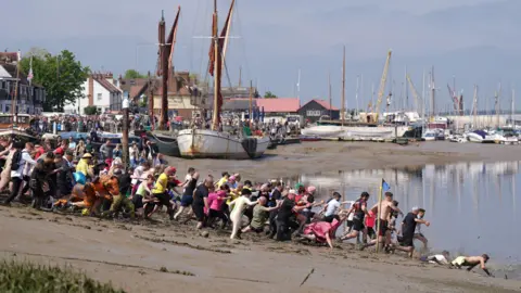 PA Media Hundreds of people, many in fancy dress, are running through mud into Hythe Quay in Maldon. There are boats in the background and historic buildings.