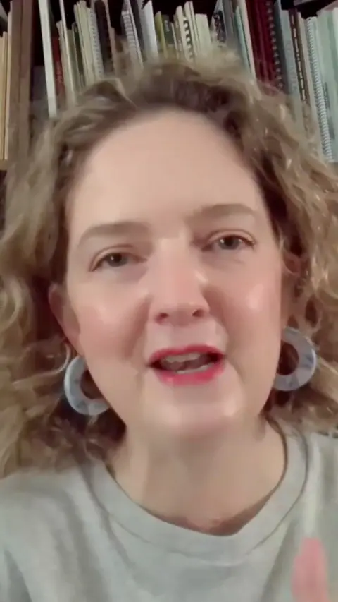 Frome and East Somerset MP Anna Sabine. She has shoulder length blonde-brown hair. She is wearing pink lipstick, a light green t-shirt and thick hooped earrings. There are books on a shelf behind her.