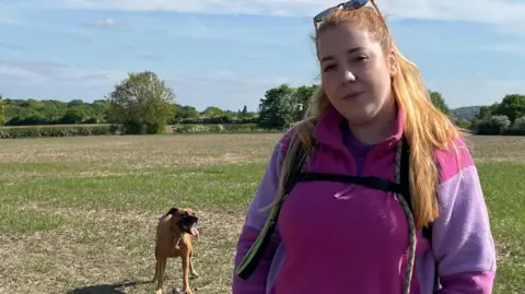 Tom MacDougall/BBC A woman with long, fair hair and sunglasses on her hear wears a pink and purple long sleeved top. She is standing in a field with tress in the distance. A brown and black dog is standing to her left.