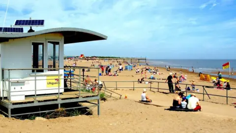 Getty Images A white RNLI Lifeguards hut can be seen on the left with small solar panels on top on a raised railing with a yellow sign and flag icon saying lifeguards. Next to it is a beach and pier in the distance with several people relaxing on the beach. A red and yellow flag with an RNLI lifeguard sheet and yellow rescue equipment can be seen on the beach.