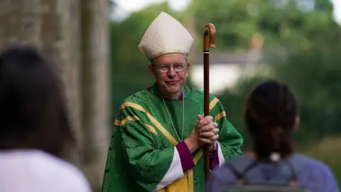 Getty Images The Bishop of Durham, Paul Butler, speaking with members of the congregation after presiding over a Eucharist service in July 2020. He is wearing a green gown with gold detailing, and carrying a wooden stick. He has grey hair and glasses.