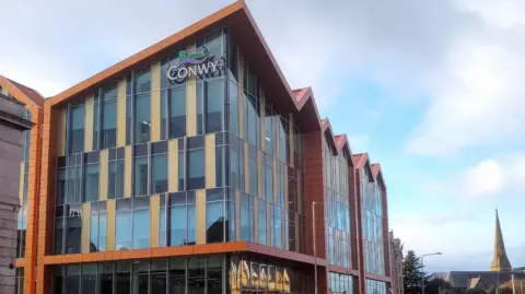 A general view of Conwy council headquarters. The four-storey building has a glass front with beige panels and the council logo at the top. 