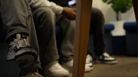 A close-up view of three people sitting indoors, showing only their legs and shoes. They wear casual jeans and trainers, and part of a wooden table leg is visible in the foreground.
