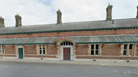 The brick-built front of Ulverston station with an empty road in front. The single-storey station has multiple chimneys, windows and doors. It has a lower roof and a second angled roof behind. It is a long building. It has a door painted green and red in the middle. 