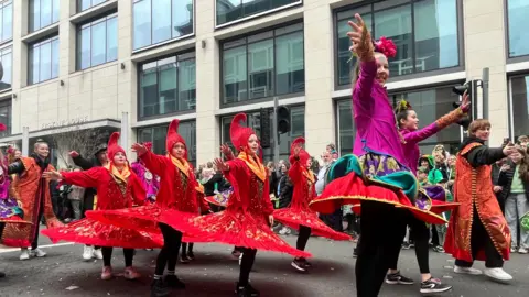 A lively street parade with dancers that wear lively costumes. Some are dressed in red outfits with golden accents, while others wear purple and multicolored costumes. Her skirts flee and show their energetic performance. The background offers a modern building with large windows and an audience that observes the celebrations.