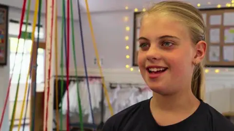 Alex Moss/BBC A smiling girl with blonde hair pulled back into a ponytail. In the background is a pole with colourful ribbons hanging from it. 