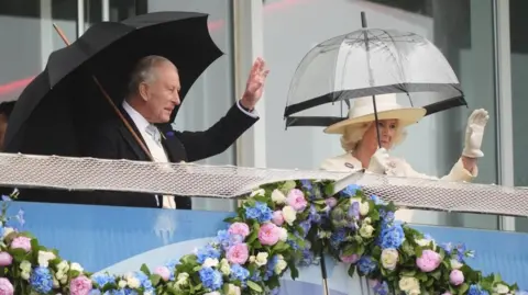 PA Media King Charles III and Queen Camilla wave to the crowd from the royal box during Derby Day at Epsom Downs.