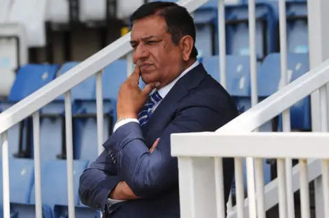 Raj Singh. He is standing in the stands of the football club. He is wearing a blue suit and is stroking his chin with one hand.