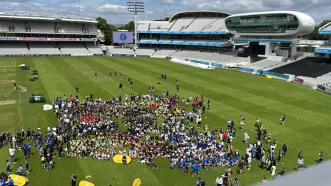 A large group of young people stand and sit on the grass at Lord's cricket ground. The stands are in the background.