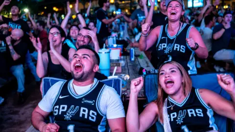 People sitting outdoors face the camera wearing Spurs jerseys looking animated and smiling while cheering