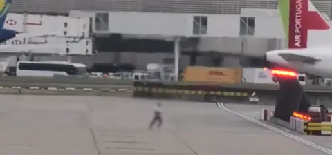 Man runs across Heathrow taxiway Screen grab from Jet TV shows a blurry image of a man running towards the tailend of a white aeroplane with vans in the background.