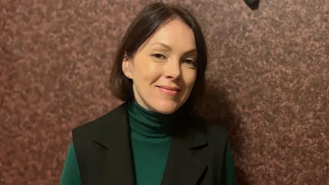 Nadia Tikhonova wearing a black sleeveless jacket with a green turtleneck jumper underneath. She is standing in front of a red patterned wall.