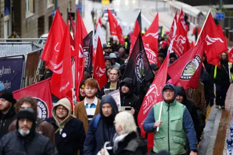 A group of people mostly men are on a city street holding banners and flags many of which are red Unite the union flags