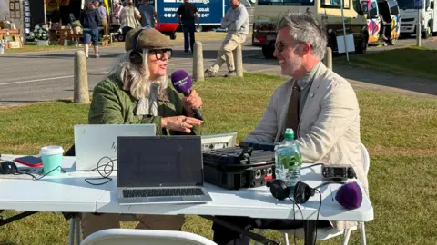 BBC/DECLAN VINK A picture of a radio presenter interviewing a guest at The Great Yorkshire Show. The presenter is wearing a flat cap, a green jacket and is holding a microphone. The guest is laughing and wearing a pale jacket. They are sitting at a table in front of an ice cream van.