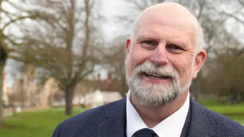 Ant Saddington/BBC The mayor of Bedford Tom Wootton is standing outdoors in a park-like setting with green grass, scattered flowers, and leafless trees in the background. They are wearing a dark textured overcoat, a collared white shirt, and a dark tie featuring a small embroidered emblem. Buildings can be seen in the distance through the trees, and the lighting suggests an overcast day.