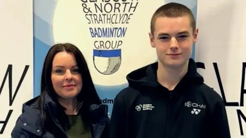 Michelle Junnor Michelle Junnor has long dark brown hair and smiles at the camera. She is wearing a green top with a black puffa jacket on top. Next to her is her son Charlie. He is taller than her and wears a black hoodie. Behind them a sign reads "Glasgow & North Strathclyde Badminton Group".