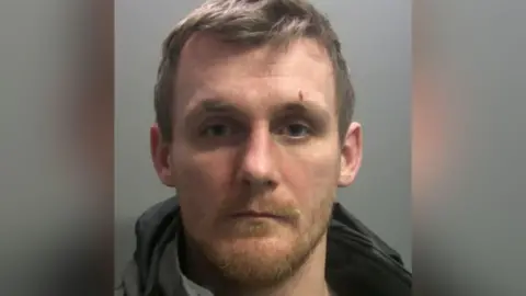 A mugshot of Marc Proudfoot, a man in his 30s with short hair and stubble. He appears to have a marking above one of his eyebrows - it is not clear whether it is a wound.
