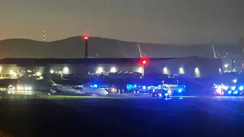 BBC A long-range shot at night-time shows an airplane on a runway surrounded by blue lights of various emergency service vehicles