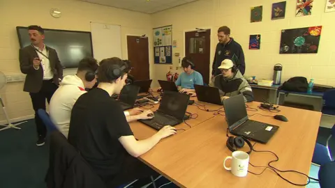 Five teenagers sitting at a desk playing a video game on laptops, with two male teachers watching on