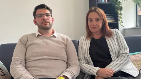 The two parents are looking straight into the camera. Michael has a cream jumper on whilst Kirsty has a striped suit jacket on an black top. They are sitting on a sofa with a book case in the background and some plants.