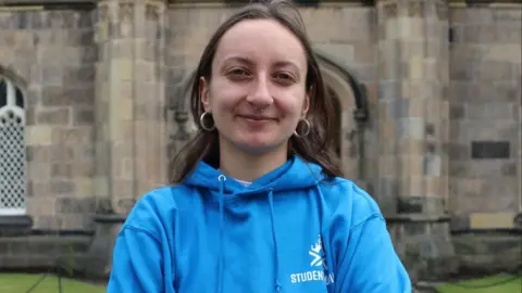 Christina Schmid, a woman with long brown hair, a blue hoodie and nose piercings, looks into the camera