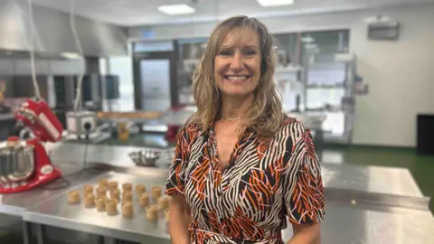 Kate Bradbrook/BBC Rachel Roy is standing in an industrial kitchen. She has long fair hair and is wearing an orange, white and black patterned dress.