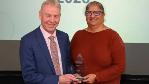 North Yorkshire Council Ms Kaur stands side by side with North Yorkshire's Council's CEO Richard Flinton, receiving her award at North Yorkshire’s Community Awards. They both hold a side of a diamond-shaped glass award in their hands. Mr Flinton has short white hair and wears a pink shirt, pink patterned tie and a blue suit jacket. Mr Kaur has grey hair worn up, and wears black-framed glasses and a red long sleeved top.