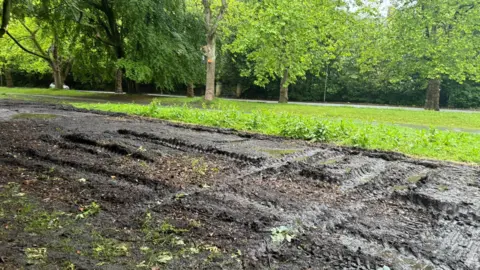 A thick muddy area of grass with vehicle marks on it. A green patch of grass can be seen behind.