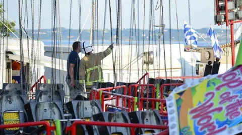 Pacemaker The men inspect a ride at a funfair One is wearing a high viz jacket and a hard hart.. The sea is in the distance. 