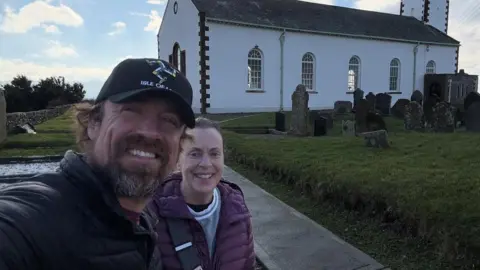 Reed Timmer on the left, has dark hair and a beard and is wearing a black coat and black cap with the Three Legs of Man emblem on it. Next to him to the right is his mum smiling, wearing a purple jacket and her dark hair tied back. They're standing in a churchyard with grave stones and a white church building behind them to the right on a sunny day.