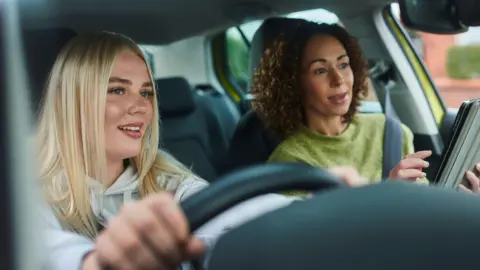 Getty Images A young blonde woman wearing a white hooded jumper appears to be taking a driving lesson, and is taking instruction from an older, but still quite young woman, who has dark curly hair and a woolen green jumper. They both look like they are looking at something in front of the car 