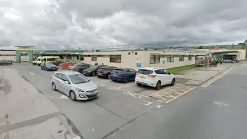 A single-storey large building. Cars parked along one side with two ambulances also parked. A sign reading Outpatients over one side of the building and Emergency over the other side.