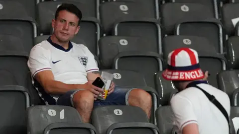 PA Media A spectator in a white England shirt and denim shorts sits alone in the stands looking dejected. Another person can be seen on a lower row to his right, wearing a white shirt and England bucket hat