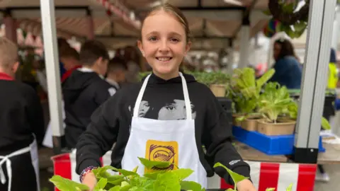 Isla-Rose wearing a black hoodie and a white apron over the top. She has her hair tied back and his holding a tray with plants in.