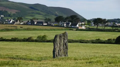 Culture Vannin A large rock is in the middle of a field, you can see a village and hills in the background.