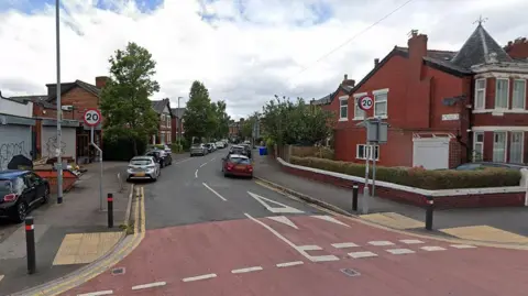 Nicolas Road, Chorlton, showing a car parked on the pavement and a skip filled with rubbish on the left, with 20mph speed limit signs and parked cars lining the road of houses.