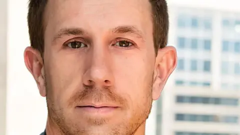 Close-up photo of Michael Forbes looking at the camera. He has short hair and a stubble.