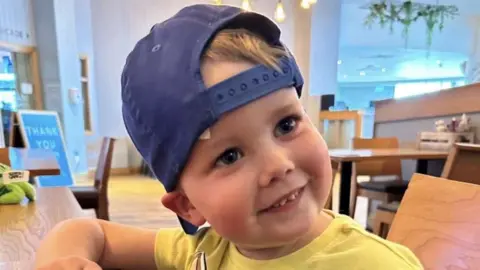 Elias is seen smiling at the camera with a cap worn backwards and wearing a yellow t shirt. 