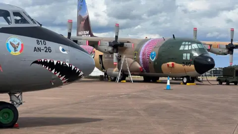 Two planes, one has a shark face painted on its nose, the other is green, brown and pink